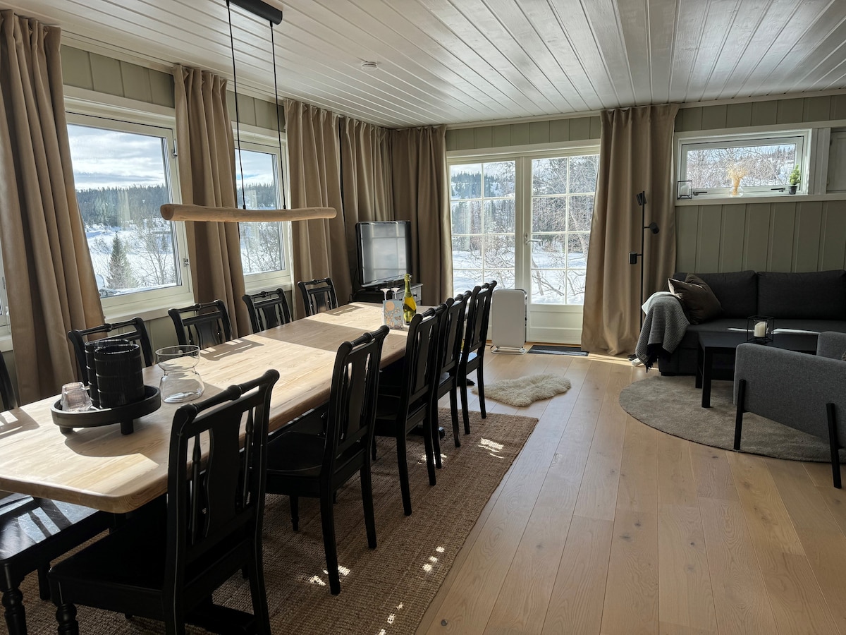 A spacious dining area is furnished with a long wooden table surrounded by black chairs. Large windows offer natural light and views of the landscape. A cozy seating area is present with a dark couch, and light curtains frame the windows.