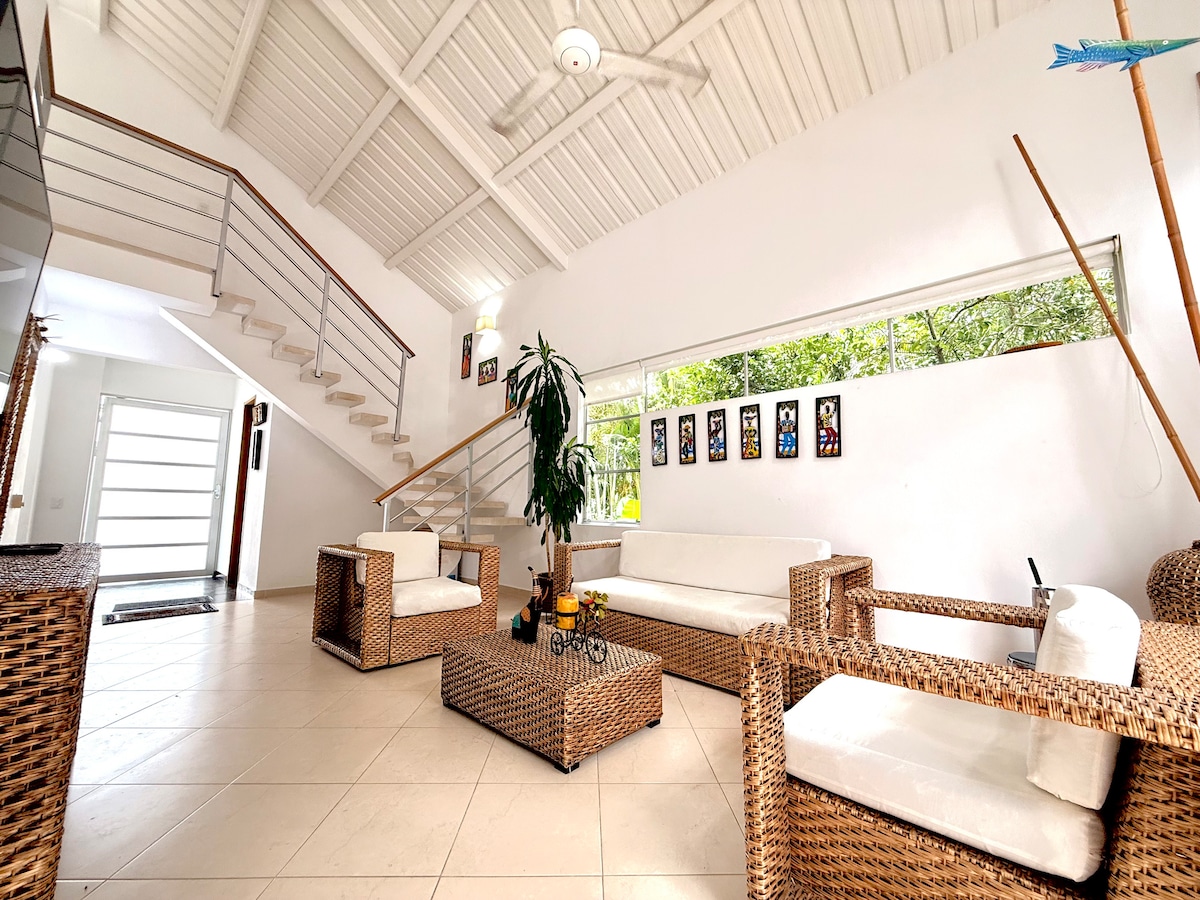 An airy living space is presented with a high, sloped white ceiling and large windows allowing natural light. Woven furniture, including a spacious sofa and armchairs, is arranged around a central coffee table. A staircase is visible in the background, enhancing the open feel of the room.