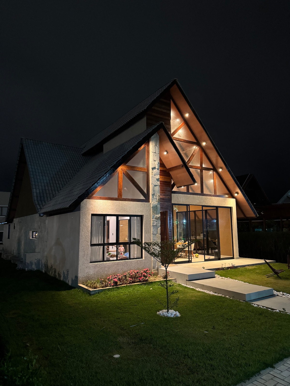 A newly constructed chalet is showcased at night, with a distinct a-frame design and large glass windows. Soft exterior lighting highlights the architectural features and manicured lawn, while a pathway and shrubbery lead to the entrance, inviting guests to explore.
