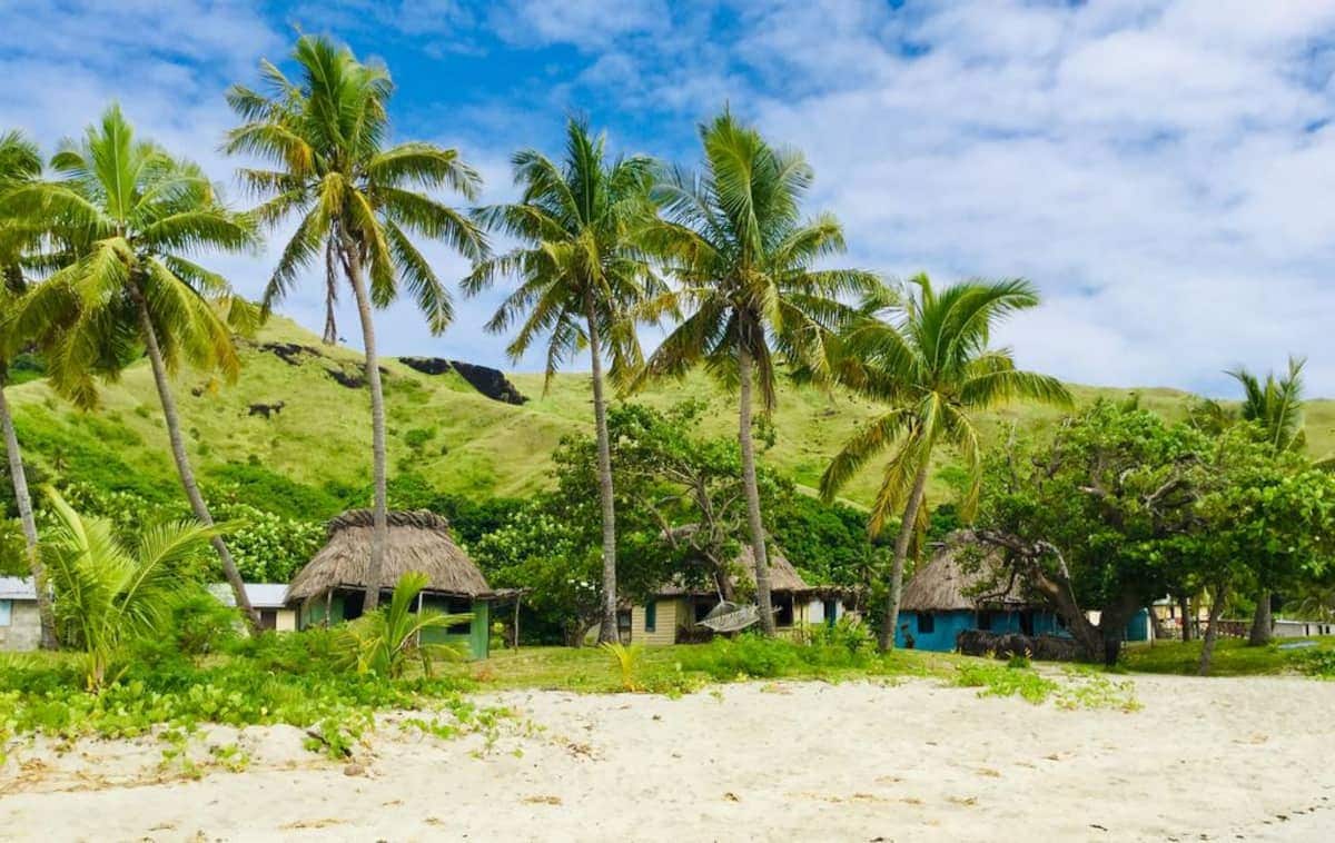Palm trees sway gently alongside traditional thatched-roof structures set against a backdrop of lush green hills. Soft white sand stretches in the foreground, while the sky displays a mix of clouds and blue, creating a serene tropical environment.