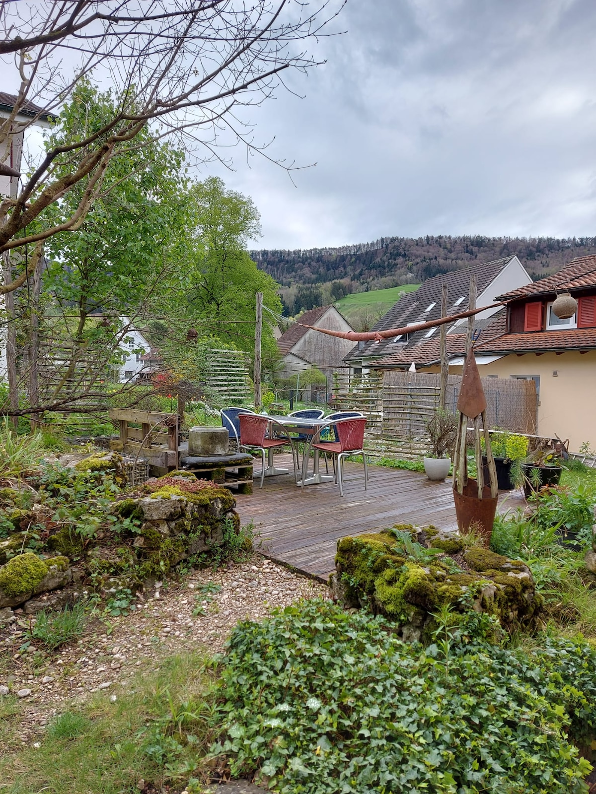 A wooden deck is visible, furnished with a table and chairs, surrounded by lush greenery. Rolling hills and tree-covered slopes can be seen in the background, while garden features, including stones and planters, add to the natural setting.