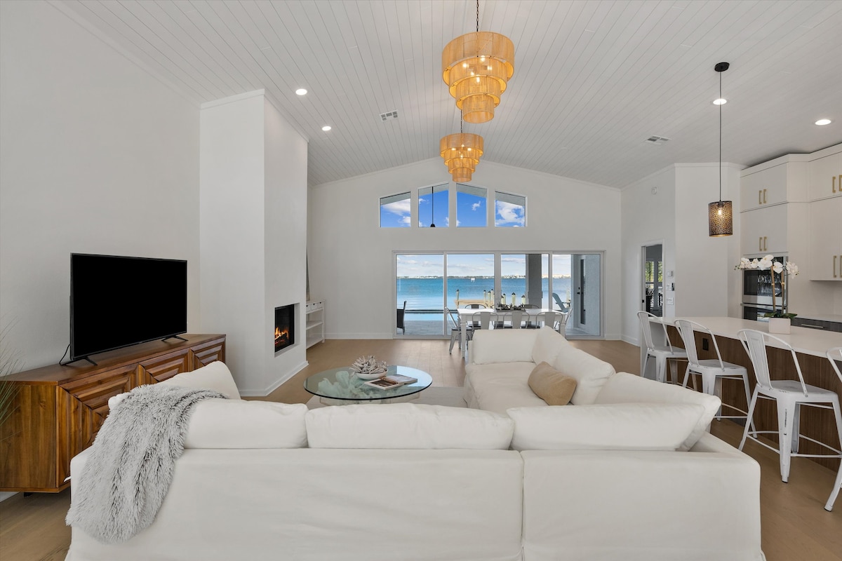 A spacious living area features a white sectional sofa facing a flat-screen television. A glass coffee table sits in front, and large windows offer a view of the water outside. Bright light fills the room, highlighting the modern decor and open layout.