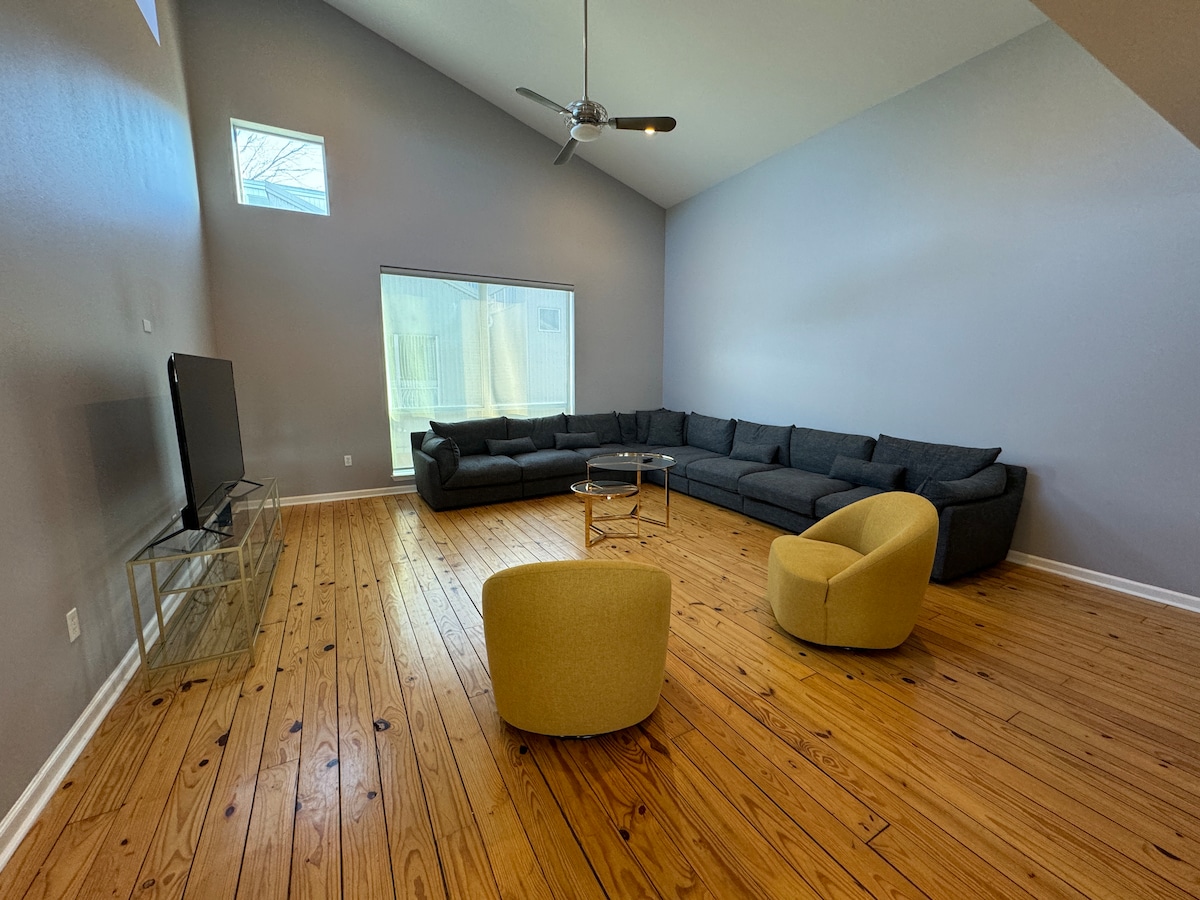 A spacious entertainment area features a large sectional sofa arranged in an inviting manner, complemented by two accent chairs. A modern coffee table sits at the center. Sunlight filters through a large window, illuminating the warm wooden flooring.