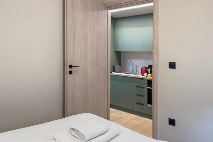 Acogedor dormitorio con vistas a una cocina moderna con gabinetes verdes mate y detalles de madera. Un diseño cómodo y funcional ideal para una vida compacta.