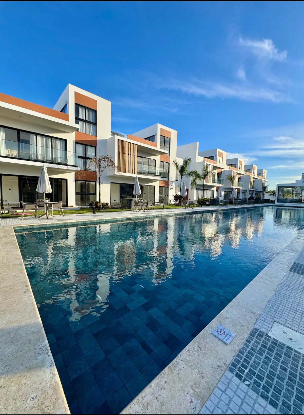 The image displays a serene swimming pool reflecting nearby modern residential buildings. Sun loungers with umbrellas are positioned along the poolside, and lush palm trees provide greenery. A clear blue sky enhances the spacious outdoor area, inviting relaxation and leisure.