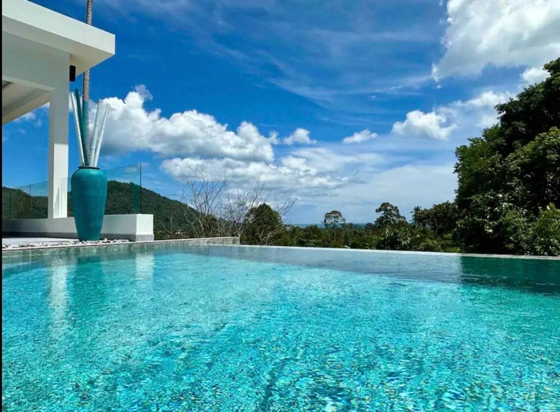 A sparkling infinity pool is showcased, reflecting the clear blue sky and lush greenery surrounding the villa. A large turquoise vase adds a vibrant touch near the pool's edge, enhancing the tranquil setting.