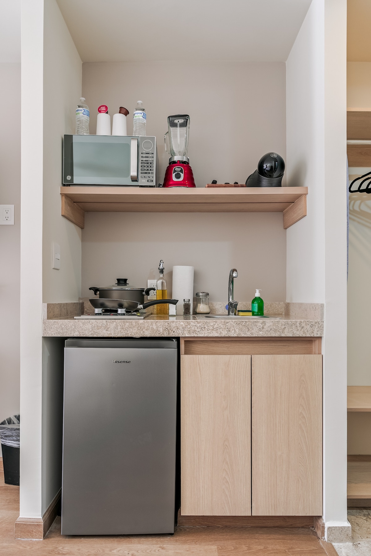 The compact kitchen area features a stainless steel refrigerator, a microwave, and a blender placed on a wooden shelf. A coffee maker and a range of condiments are visible, along with a sink and cooking supplies organized on the countertop.