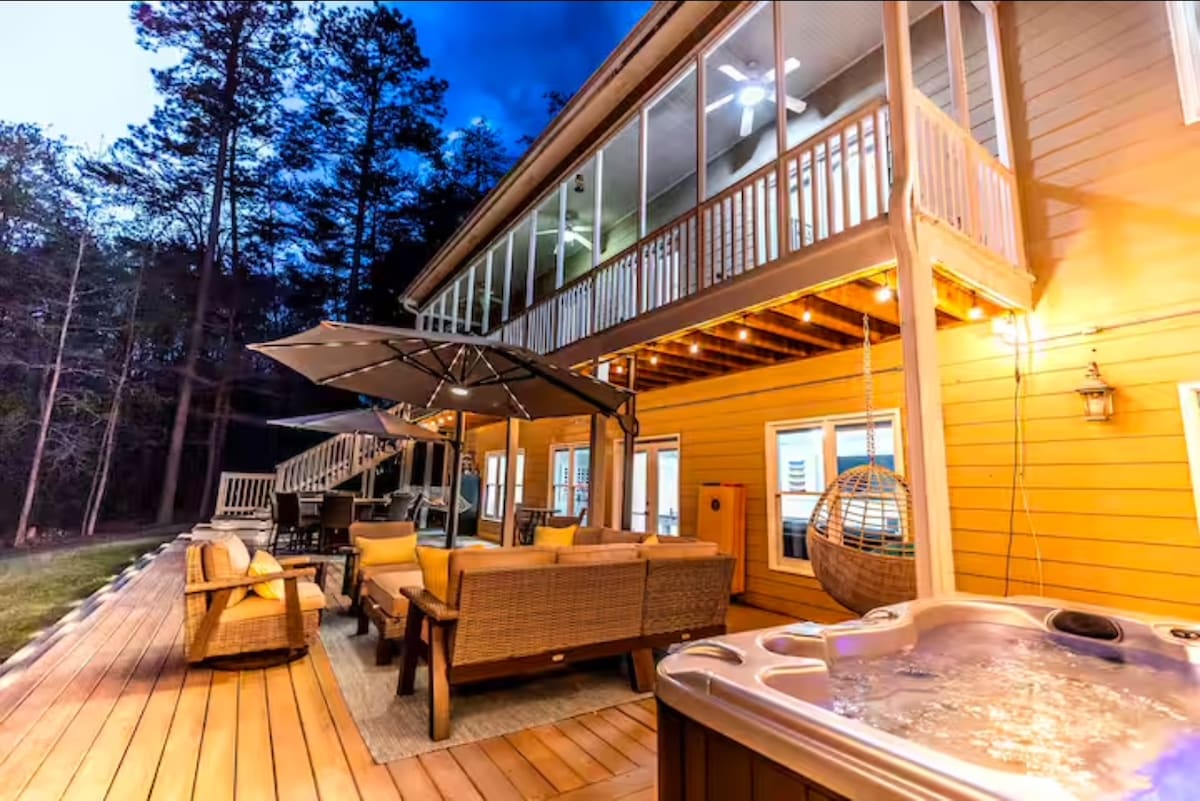 A spacious outdoor area features comfortable seating arrangements with wicker furniture and a large umbrella. Soft lighting highlights the wooden deck, and a hot tub is positioned nearby, providing a cozy place to relax. The backdrop includes a two-story house with numerous windows.