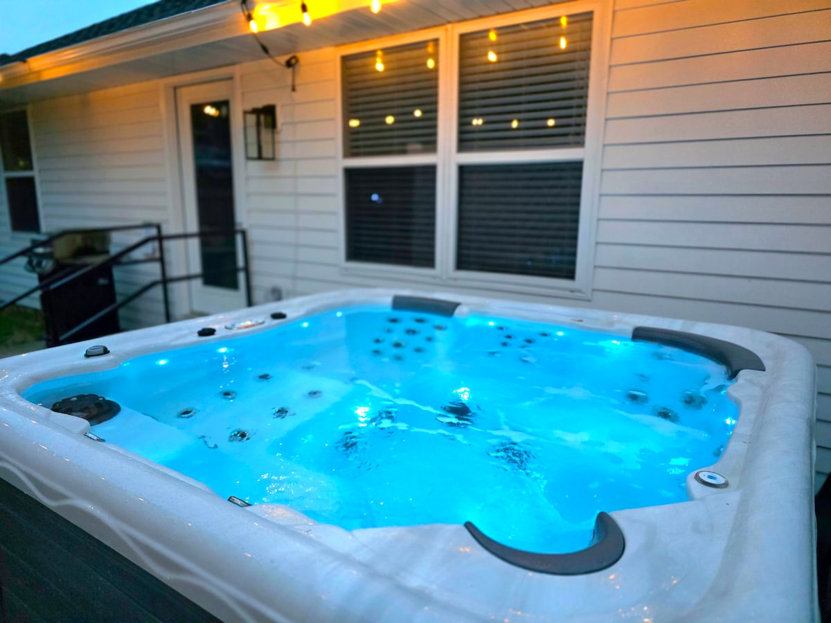 A private hot tub sits outdoors, illuminated with soft blue lighting. The surround features a clean, modern design, with windows reflecting a warm glow from string lights above. The space is ideal for relaxation, creating a serene ambiance for evening unwinding.