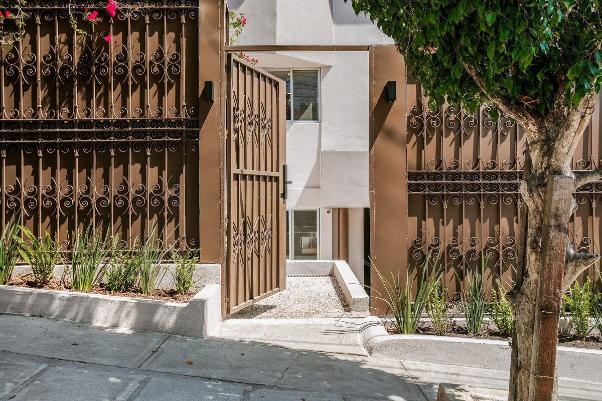 An elegant entrance features decorative metal gates, framed by lush green plants and trees. The pathway is lined with smooth stones, leading to a well-lit interior, suggesting a welcoming ambiance. The contemporary building design is highlighted by neutral-colored walls.