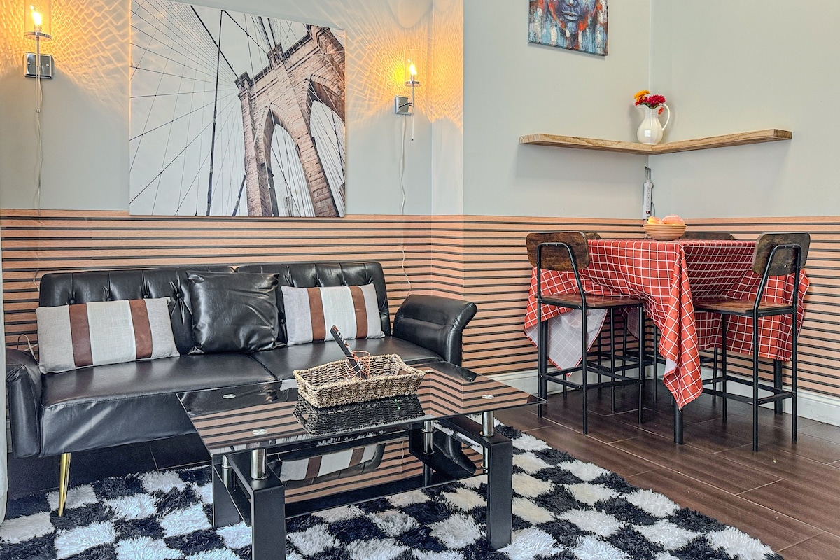 A stylish living area features a black leather sofa with contrasting cushions and a sleek glass coffee table. A cozy dining table covered with a red checkered tablecloth is positioned beside an elegant wall shelf. Artwork and ambient lighting enhance the modern design.