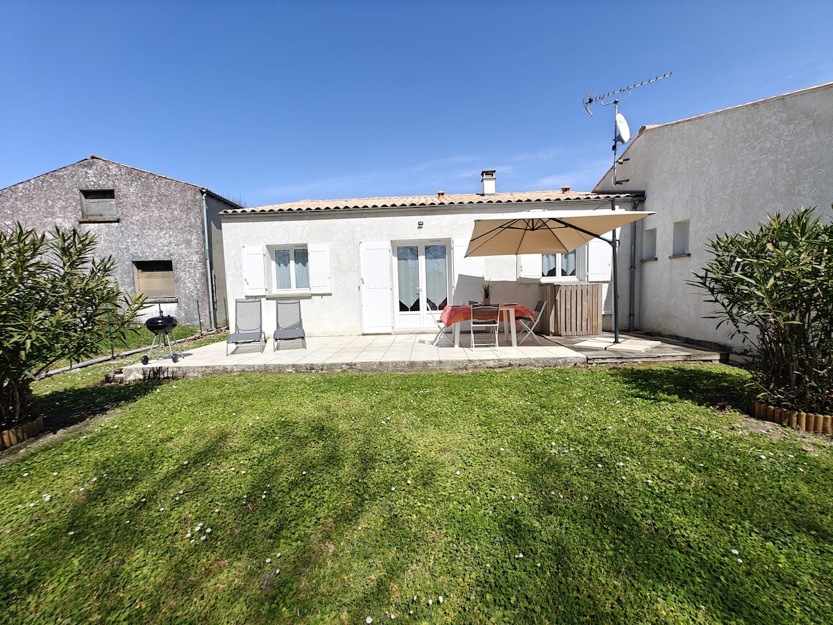 A private garden is featured, framed by low greenery and a gravel patio. A large umbrella provides shade over a table and chairs. A barbecue grill is visible, and the exterior of the house presents a clean, white facade with multiple windows and doors.