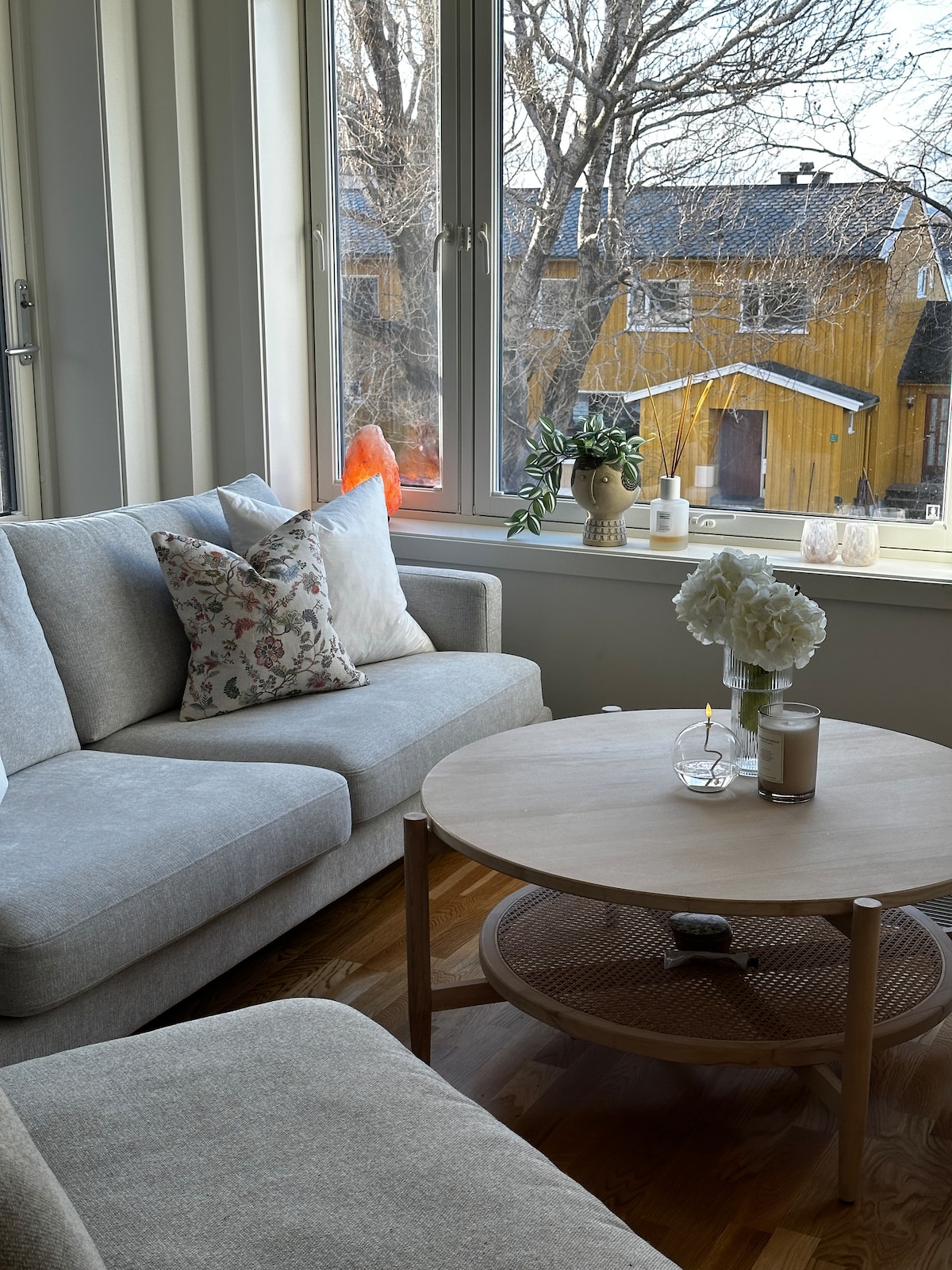 A light-filled living area features a grey sectional sofa adorned with patterned cushions. A circular wooden coffee table supports candles and a vase of white flowers. Large windows offer views of the surrounding neighborhood, framed by trees.
