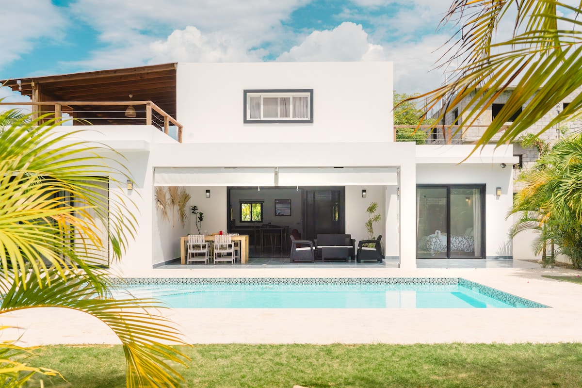A modern villa features a sleek, white exterior and an inviting outdoor space. Floor-to-ceiling glass doors open to a private pool surrounded by landscaped greenery. A shaded patio offers seating and dining options, creating a welcoming area for relaxation and entertainment.