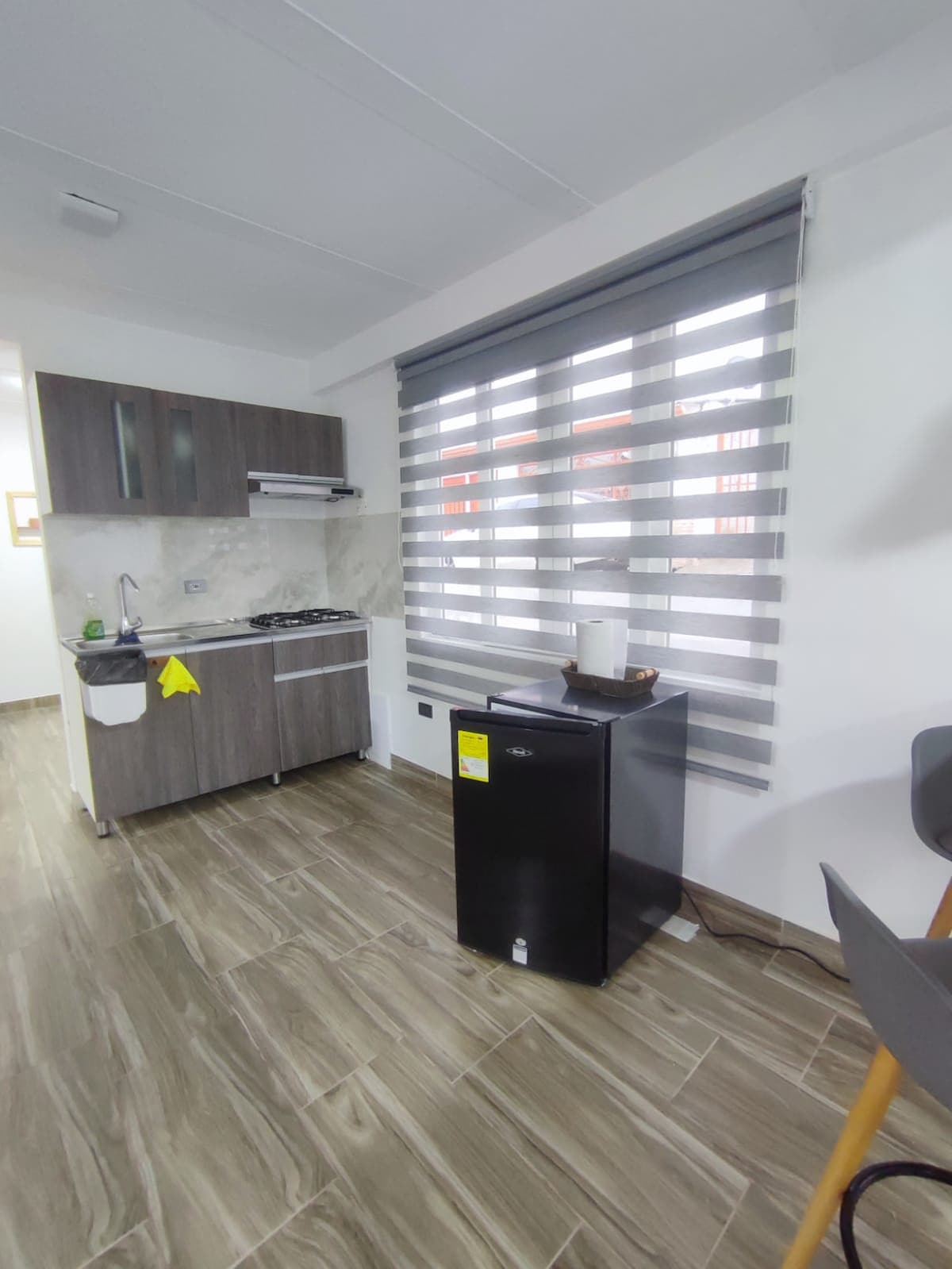 A compact kitchen area is presented with modern cabinetry and a four-burner stove. A dark refrigerator stands next to a window adorned with striped blinds, allowing soft light to filter through. A small table and chair complete the functional space.