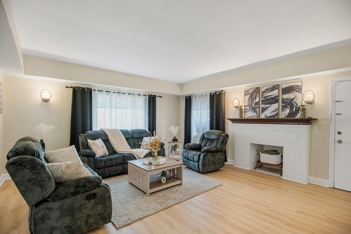 A spacious living area features plush seating arranged around a central coffee table. Soft light filters through sheer curtains, illuminating the light-colored walls and wooden flooring. Artwork adorns the wall above a white brick fireplace, creating a cozy focal point in the room.