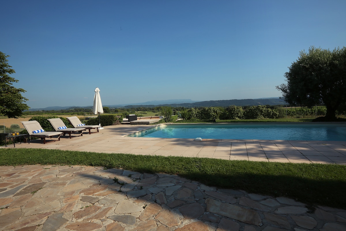 A tranquil outdoor space features a swimming pool surrounded by lounge chairs. A sun umbrella provides shade, and lush greenery borders the area. The view extends over the valley, with distant mountains visible under a clear blue sky.