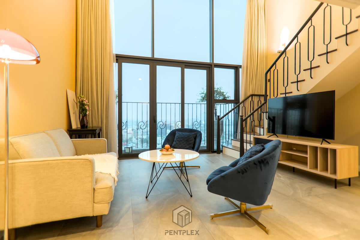 The spacious living area features large floor-to-ceiling windows that provide natural light and views of the outside. A light-colored sofa and two accent chairs create a comfortable seating arrangement. A round coffee table sits at the center, complemented by a wall-mounted smart TV.
