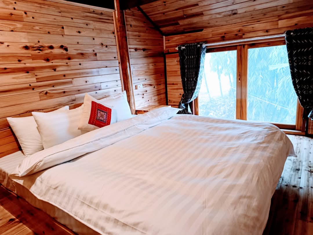 A well-appointed sleeping area features a spacious bed adorned with crisp linens and a decorative pillow. Natural wood panels create a warm ambiance, while large windows offer a serene view of the outdoor landscape, framed by light, patterned curtains.