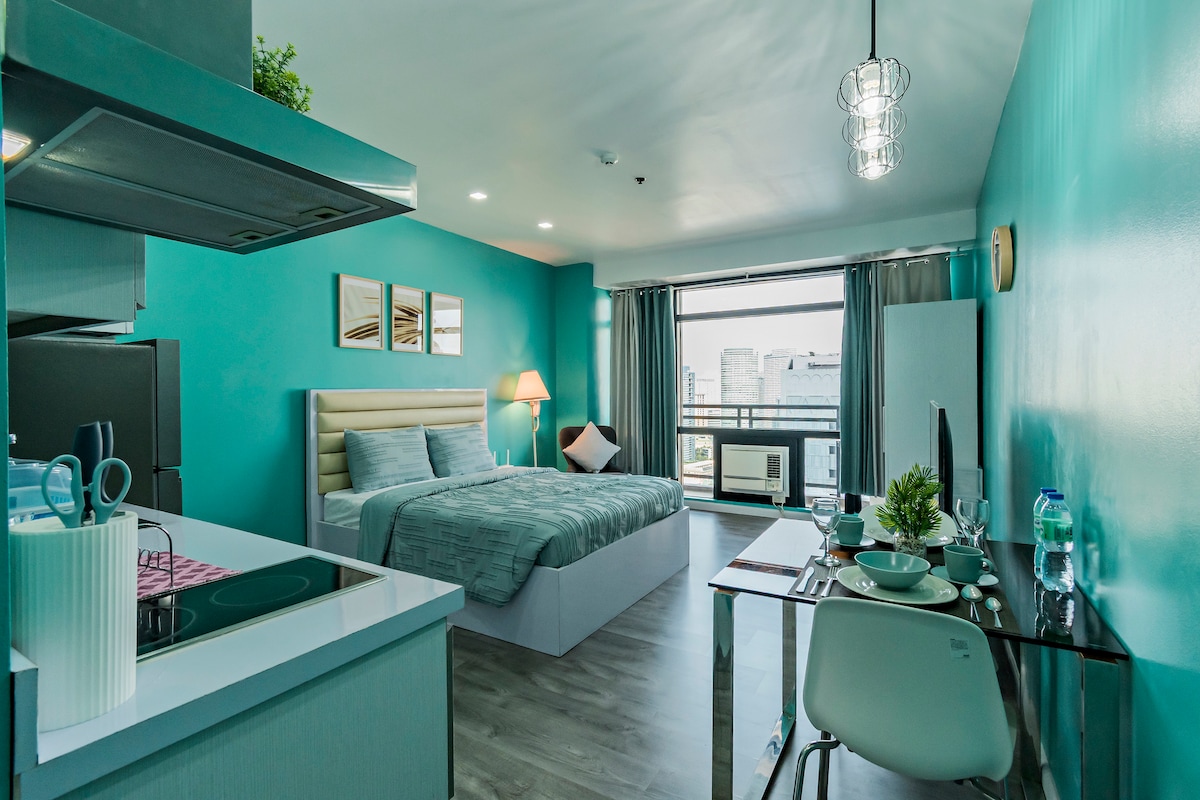 A well-appointed studio layout features a queen-sized bed with light blue bedding. A modern kitchen area is equipped with essential appliances, while a dining table for two complements the workspace. Large windows offer natural light, with city views visible beyond.