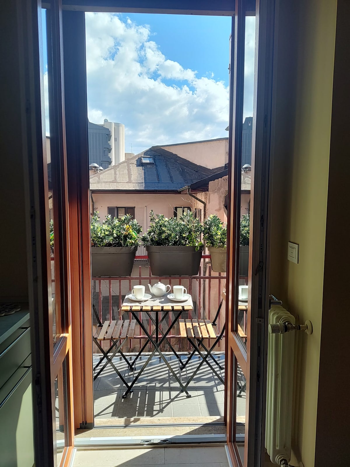 A bright balcony is showcased, featuring a small table and two chairs. Flower pots line the railing, adding greenery to the space. Sunlight streams through large, open doors, illuminating the area and highlighting the rooftops of the historic buildings in the background.