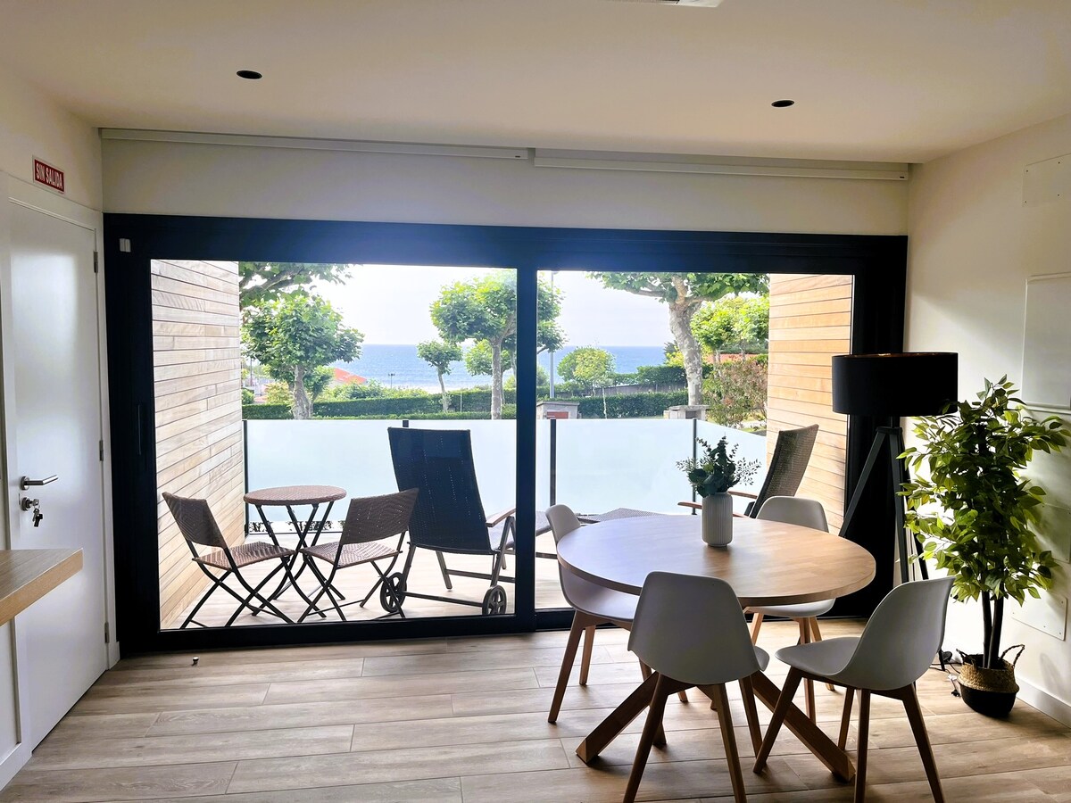 A bright living area features large glass doors that open to a private terrace. A round dining table with four chairs sits adjacent to a potted plant. Outdoor seating, consisting of two chairs and a small table, is visible beyond the glass, with a glimpse of greenery and the sea.