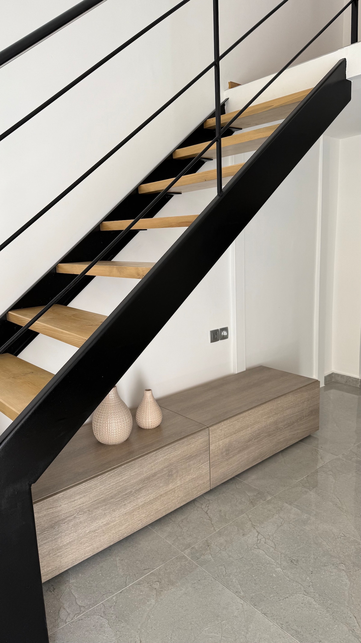 A modern staircase with wooden steps and a sleek black railing ascends to the upper level. Below, a minimalist wooden bench is positioned against the wall, accented by three decorative vases in soft beige tones.