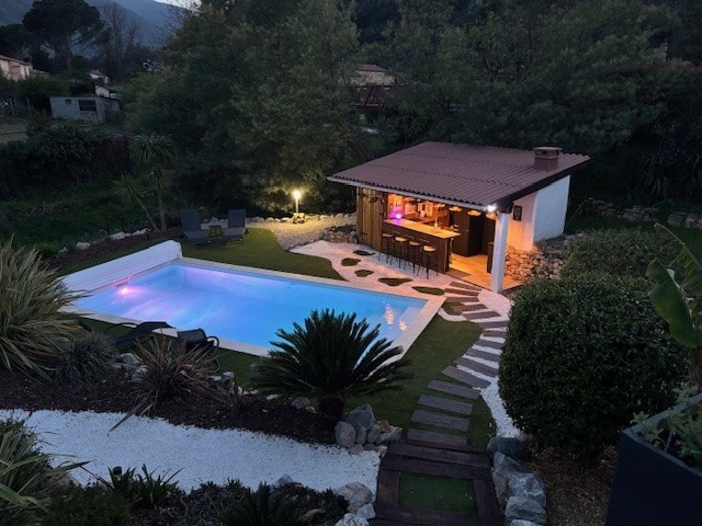 A serene outdoor area features a well-maintained pool illuminated with soft lighting. A cozy bar structure is visible nearby, surrounded by lush greenery and neatly arranged paths. Comfortable seating is positioned beside the pool, offering a peaceful retreat.