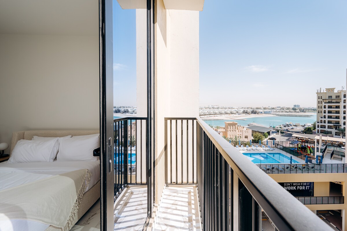 A studio apartment is shown with large glass doors leading to a private balcony, providing views of the waterfront and nearby pool area. The sleeping area features a spacious bed with white linens. Natural light illuminates the open layout, enhancing the overall ambiance.