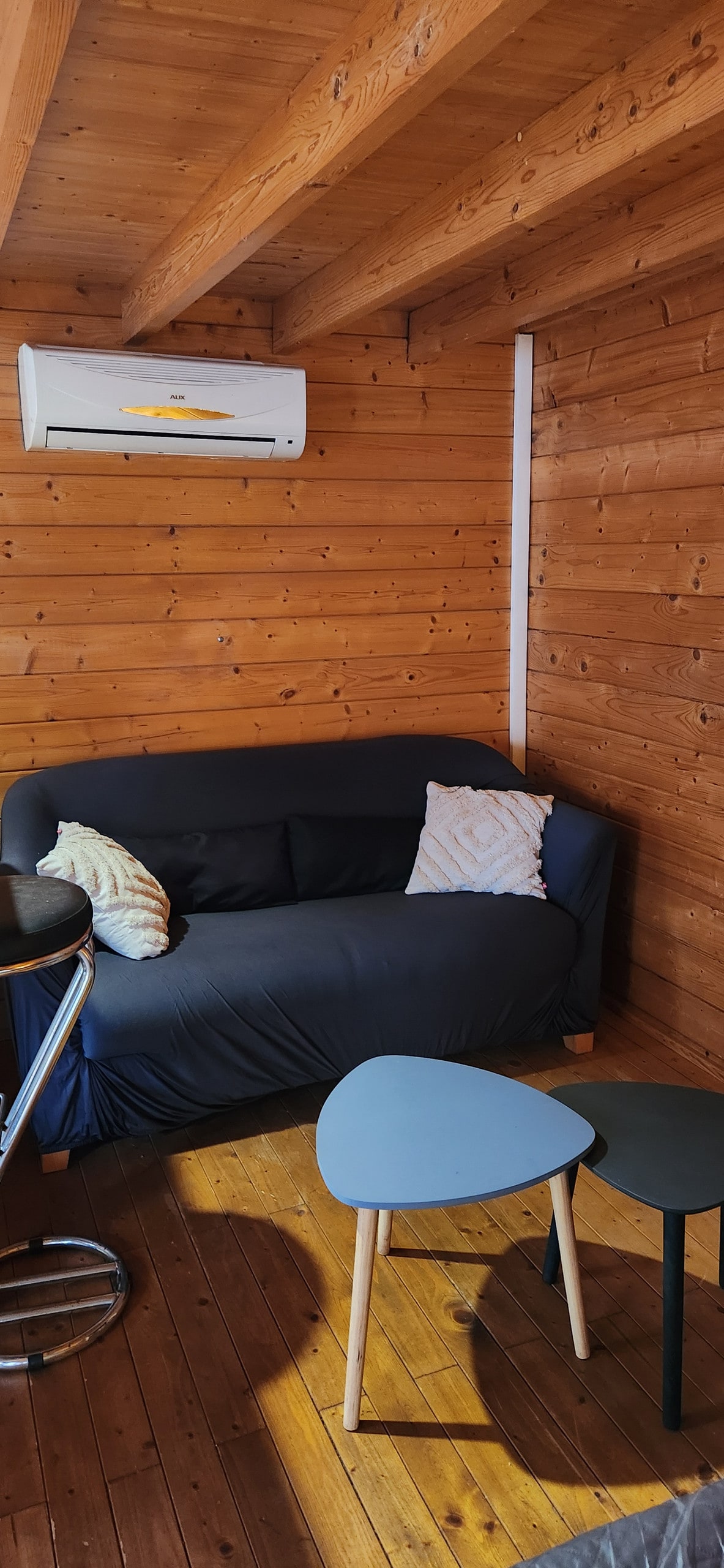 A cozy seating area features a dark sofa adorned with a decorative pillow, set against a wooden wall. Two small tables in varying shapes are positioned beside the sofa. Natural light illuminates the warm wooden floor, creating an inviting space.