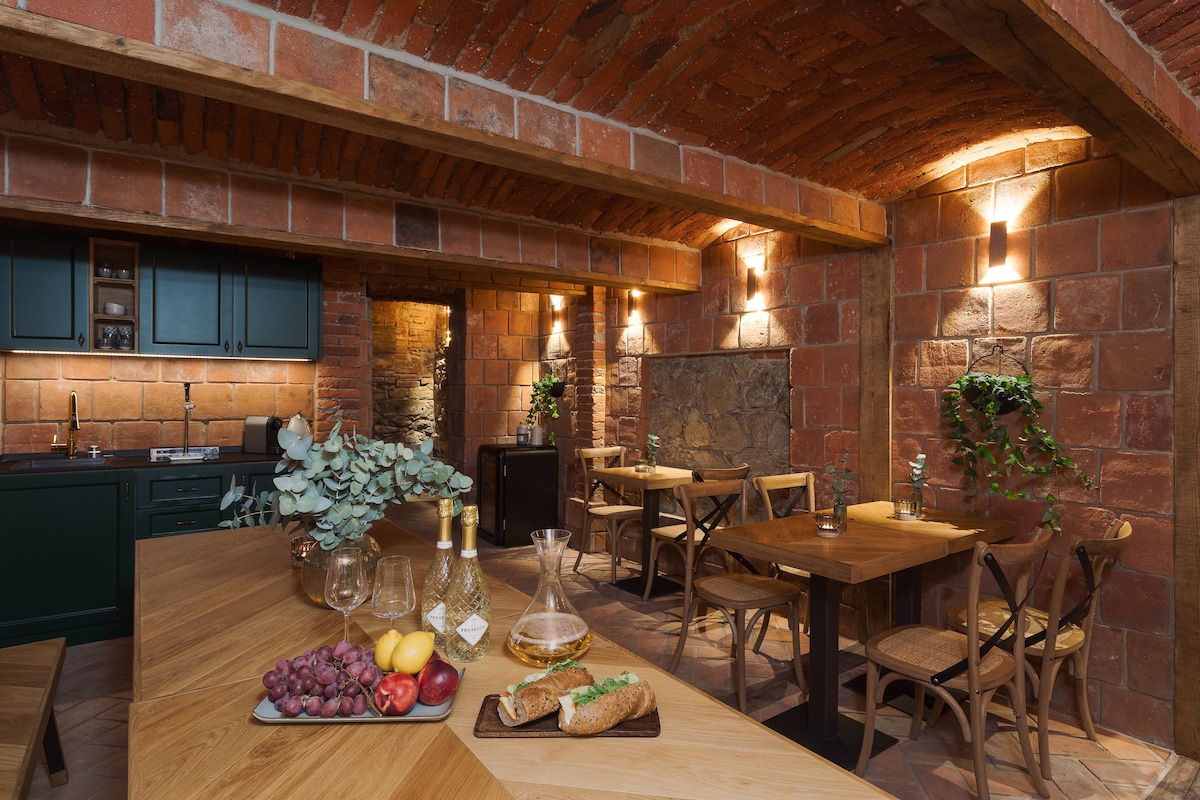 The kitchen area features exposed brick walls and wood beams, setting a rustic ambiance. A long wooden table surrounded by cross-back chairs is positioned under soft lighting. An equipped kitchen is observed in the background, complemented by natural decor elements.