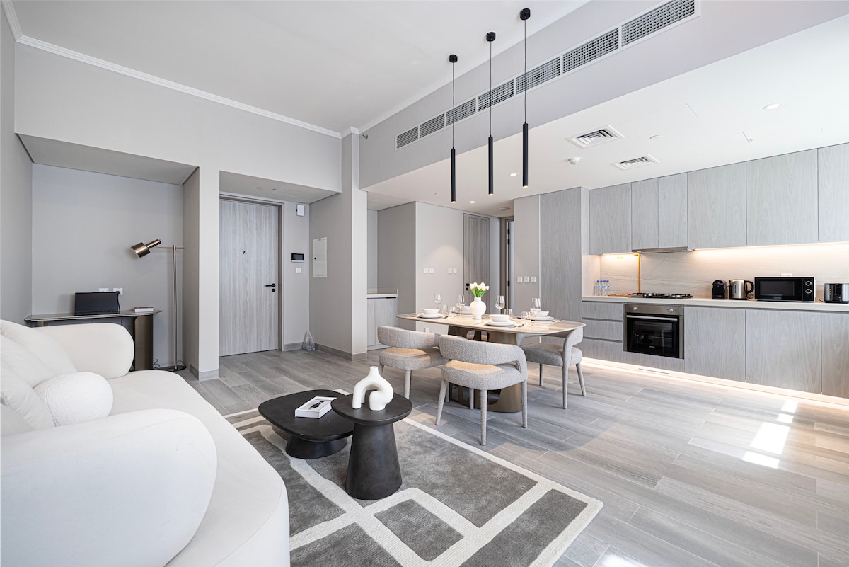 A modern open-concept living space is displayed, featuring a light-colored sofa facing a stylish coffee table. A dining area with circular seats surrounds a wooden table. Soft lighting from pendant fixtures enhances the sleek cabinetry in the kitchen, creating a harmonious blend of comfort and elegance.