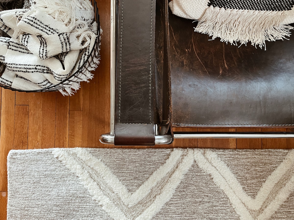 A close-up view showcases a sleek leather chair with chrome armrests, positioned beside a woven basket containing cozy, patterned blankets. The light-toned wooden floor is complemented by a textured area rug featuring a subtle, neutral pattern.