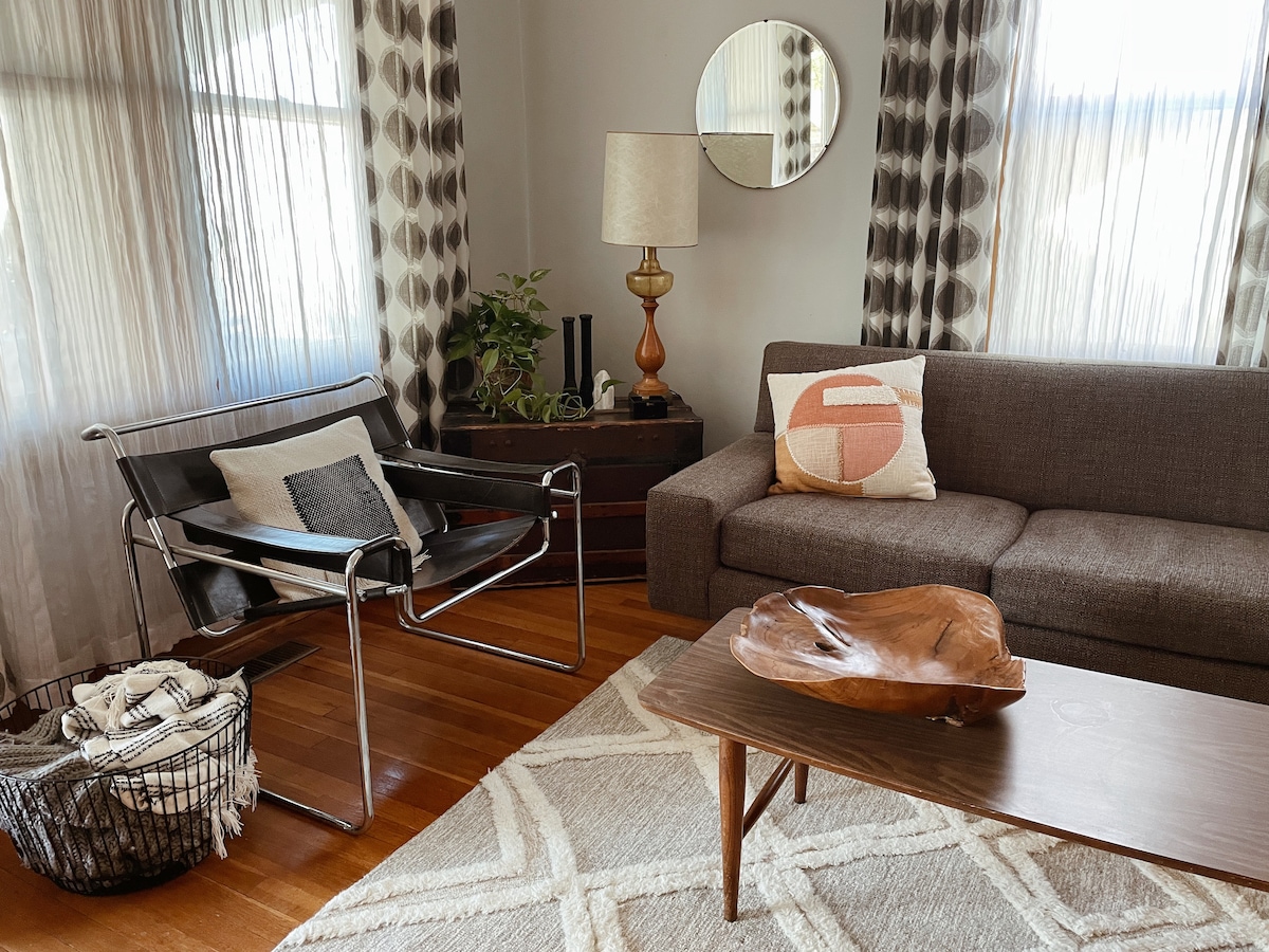 A light-filled living room showcases modern furnishings, including a gray sofa and a black metal chair. A round mirror reflects sunlight, while a wooden coffee table features an artistic bowl. Soft textiles and natural elements create a comfortable ambiance in the space.
