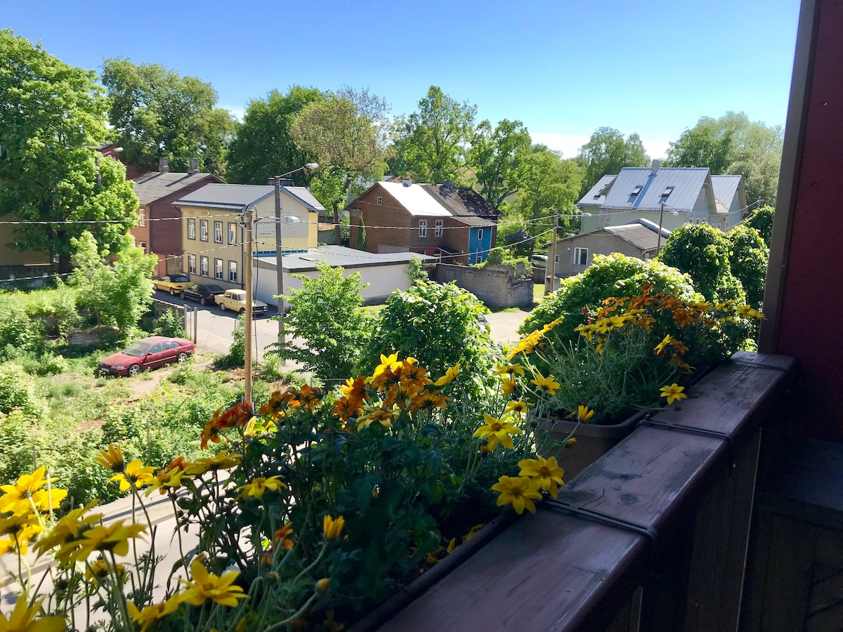 A view from a balcony showcases a vibrant flower box filled with yellow and orange blooms. Below, a neighborhood scene unfolds with trees and various buildings, including houses and garages, bathed in natural daylight.