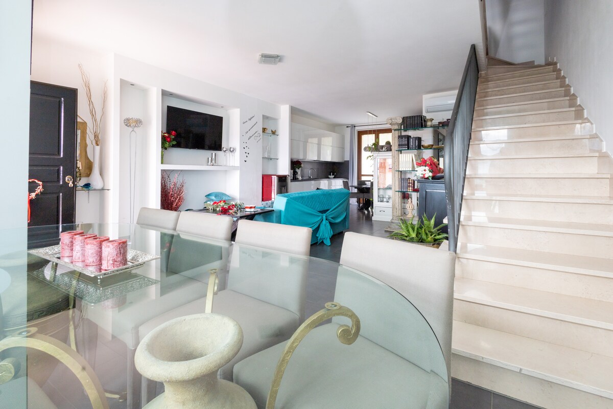 A bright and modern living area features a dining table with light-colored chairs. A contemporary kitchen is visible in the background, along with stairs leading to the upper levels. Decor elements include plants and subtle artistic touches, enhancing the open layout.