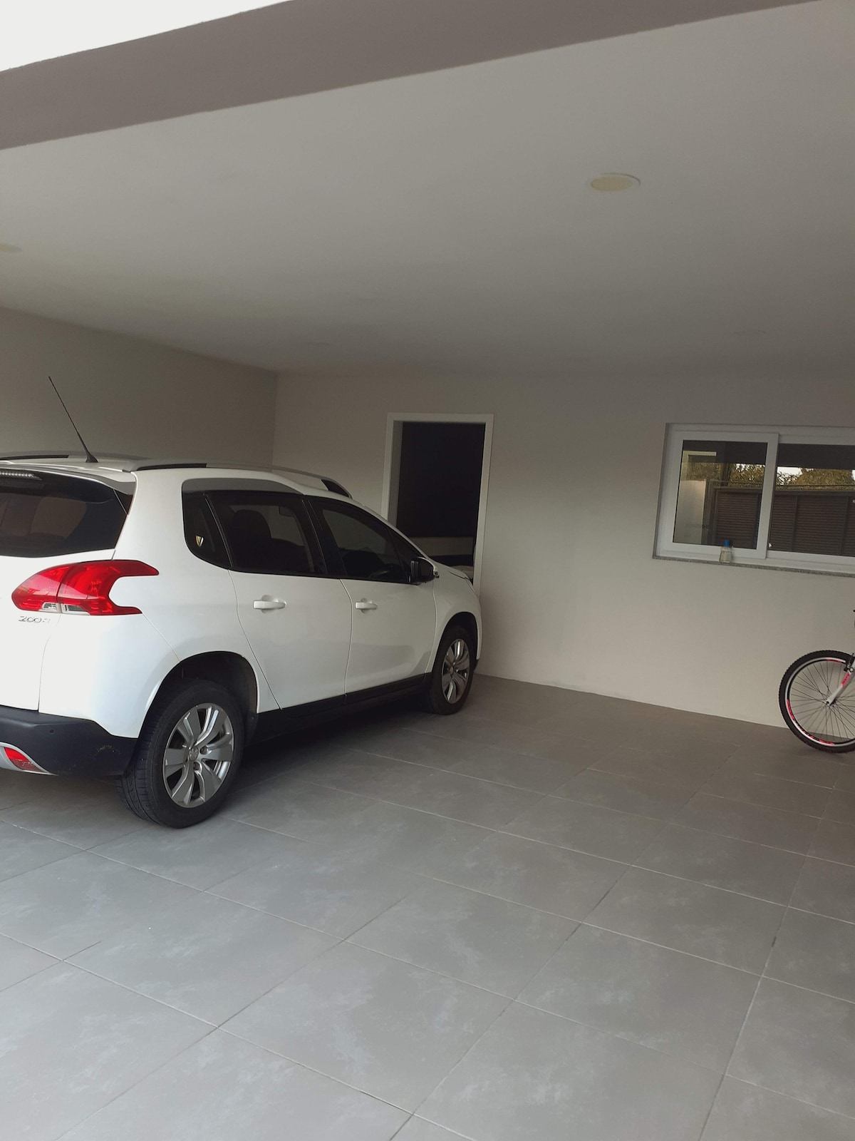 A covered parking space is depicted, showcasing a white car stationed on light-colored tiled flooring. A bicycle is positioned nearby, with an entry door and a window visible in the background, allowing natural light to enter the area.