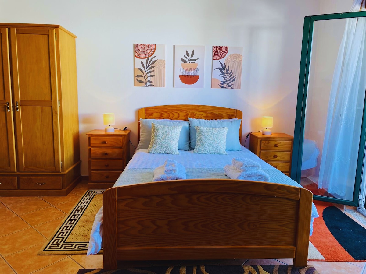 A cozy sleeping area features a wooden bed with soft blue bedding, complemented by light-colored pillows. Two bedside lamps provide gentle illumination. Framed artwork with plant motifs is displayed above the bed. A large wardrobe stands nearby, with a door open to reveal additional storage space.