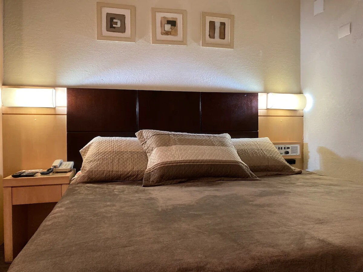 A spacious bed is set against a dark wooden headboard, complemented by soft, neutral-toned bedding and decorative wall art above. Soft lighting fixtures are mounted on both sides, enhancing the overall ambiance of the sleeping area.