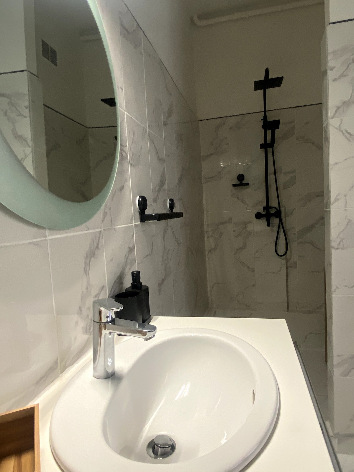 The bathroom features a minimalist design with marble-like tiles and a round mirror. A modern sink with a sleek faucet is positioned beside a black showerhead and controls mounted on the wall. The space is illuminated by natural light filtering in.