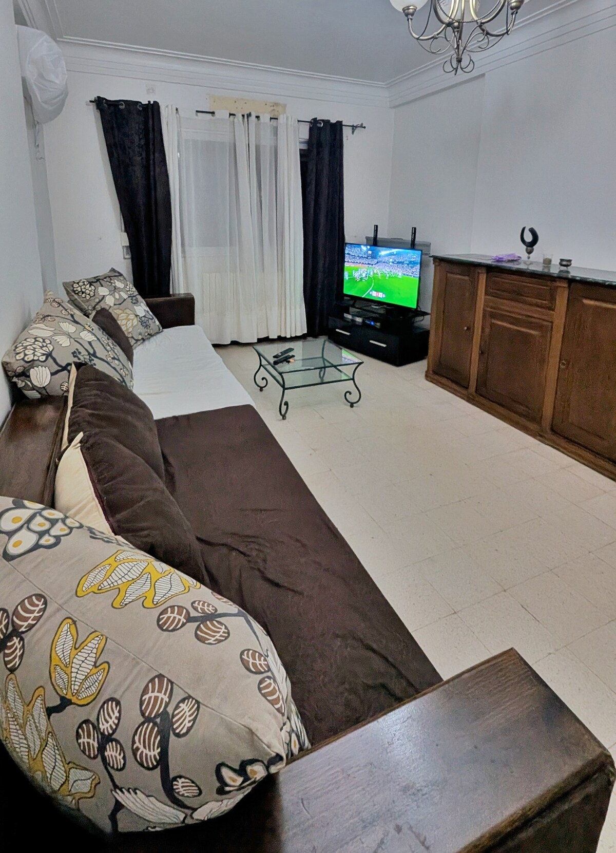 A living area is featured with a plush brown sofa adorned with decorative pillows. A glass coffee table is placed in front of the sofa, and a television with a visible screen is positioned across from it. Light-colored curtains frame a window, contributing to the bright ambiance.