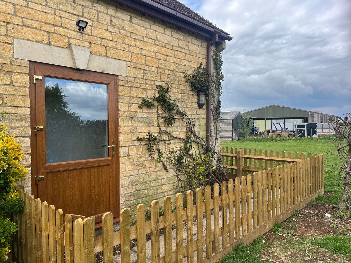 The exterior features a charming stone wall with wooden fencing surrounding a small garden area. A single wooden door with a glass panel provides entry, complemented by green foliage and a nearby farm structure visible in the background.