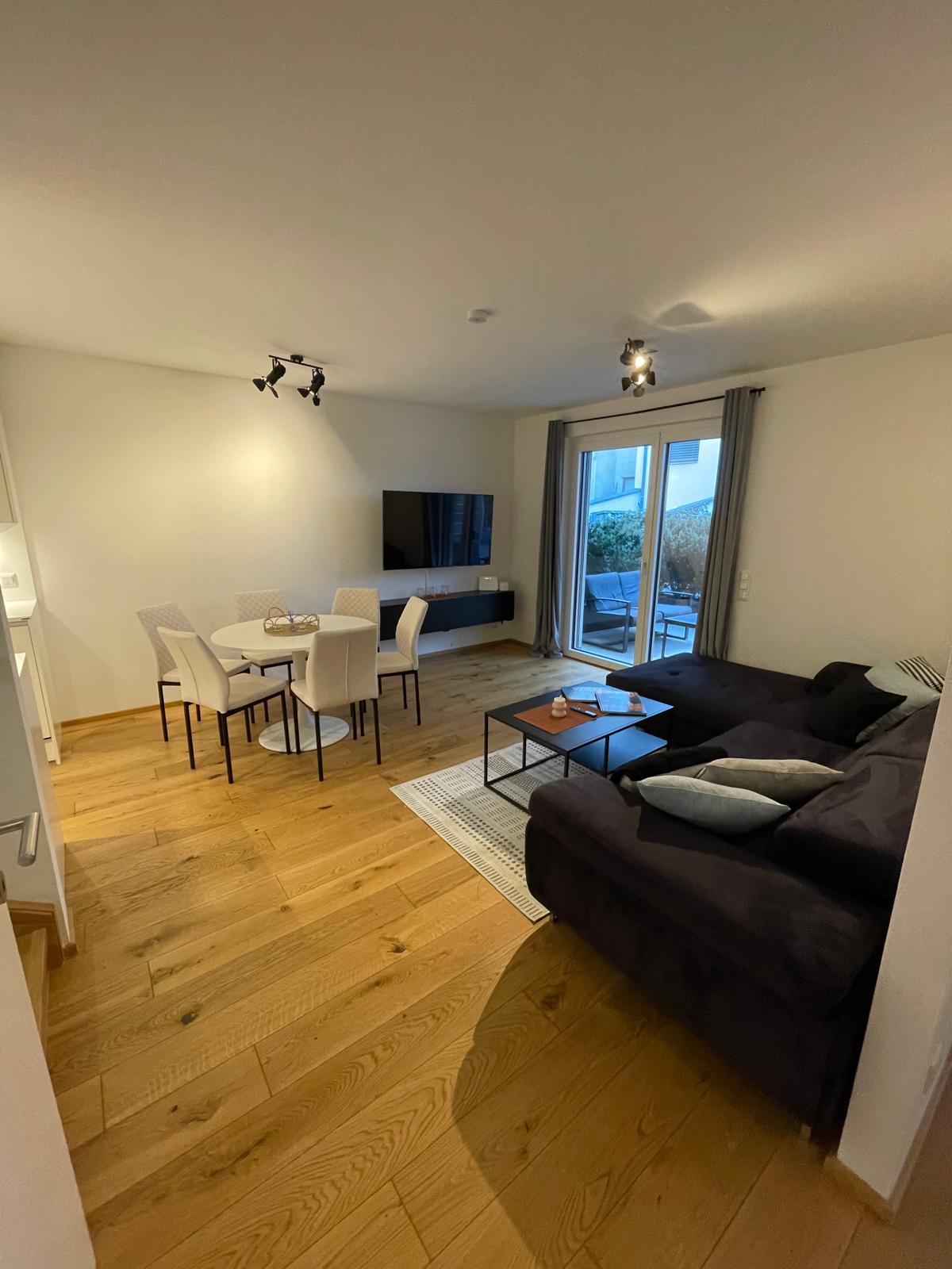 An open living and dining area features a dark sofa, a coffee table, and a dining table with six chairs. A television sits mounted on the wall, while large glass doors lead to a small outdoor space, allowing natural light to fill the room.