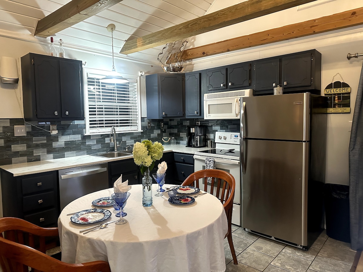 The kitchen area features dark cabinetry and tiled backsplash, complemented by a round dining table set for two. A stainless steel refrigerator and microwave are visible, along with essential appliances. Natural light enters through a window, illuminating the scene which exudes a functional, welcoming vibe.