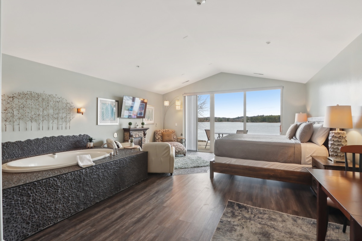 A spacious studio condo features a king sized bed beside large windows offering waterfront views. A jetted tub is situated against a textured stone wall, while a seating area includes a chair and a small table. Natural light enhances the neutral tones throughout the room.
