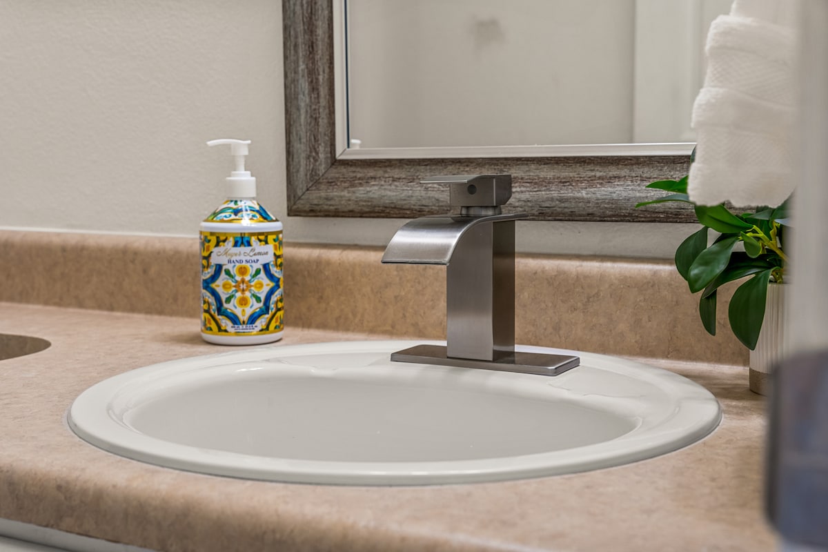 The bathroom sink area features a modern faucet and a neatly arranged soap dispenser, accented by a decorative design. A small plant adds a touch of greenery to the warm-toned countertop, while a framed mirror reflects the space.