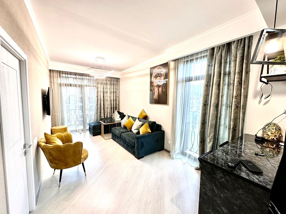 A stylish living area features a dark sofa with yellow accent cushions, accompanied by a mustard-colored armchair. Large windows are draped with light curtains, allowing natural light to fill the space. A modern artwork hangs on the wall, complementing the sleek decor.