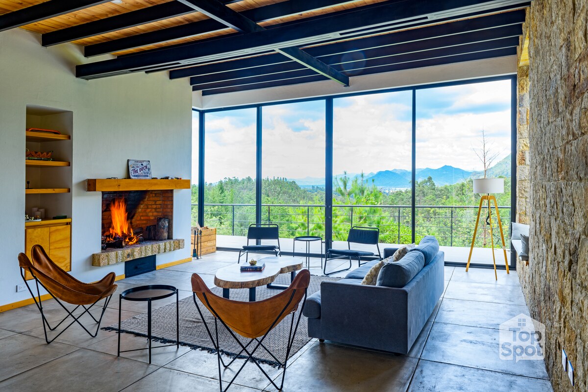 A spacious living area features large glass windows showcasing mountain views. Natural light fills the room, highlighting a comfortable sofa, two butterfly chairs, and a circular coffee table. A stone fireplace stands as a focal point, creating an inviting atmosphere.