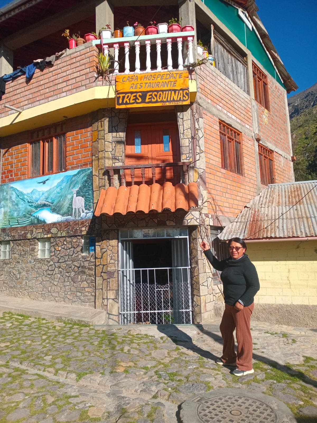 A rustic building with traditional stone and brick elements is showcased, featuring a welcoming sign for 'Tres Esquinas' above the entrance. A person stands outside, holding a branch, while colorful banners and plants adorn the upper balconies, highlighting the inviting atmosphere of the structure.