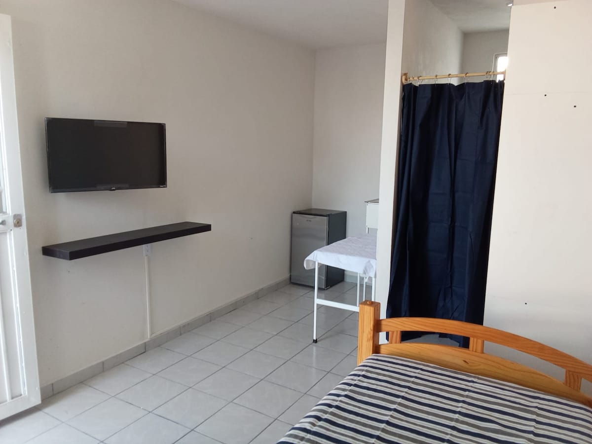 A furnished individual room is presented, featuring a single bed with a striped blanket. A wall-mounted television is visible, along with a small refrigerator and a white table. A privacy curtain separates the shower area, while tile flooring enhances the overall space.